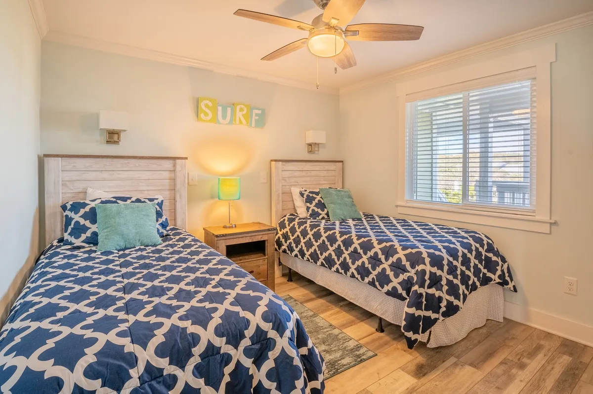 Depicts a twin bedroom with two twin beds, surf-themed decor, a colorful lamp, and a wooden nightstand. The space is bright and cheerful, creating an inviting environment for kids or friends to relax or play games.