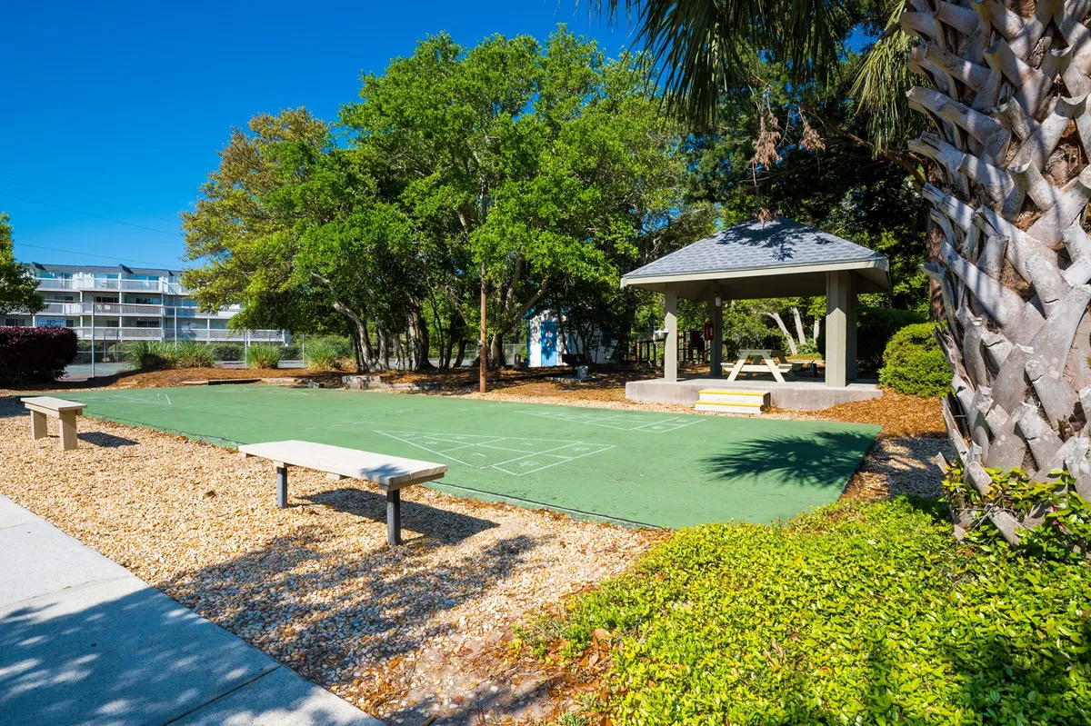 A green playing surface with a shaded gazebo, surrounded by trees and benches. The area is designed for outdoor games and gatherings, with plenty of open space for relaxing in the shade and enjoying fresh air.