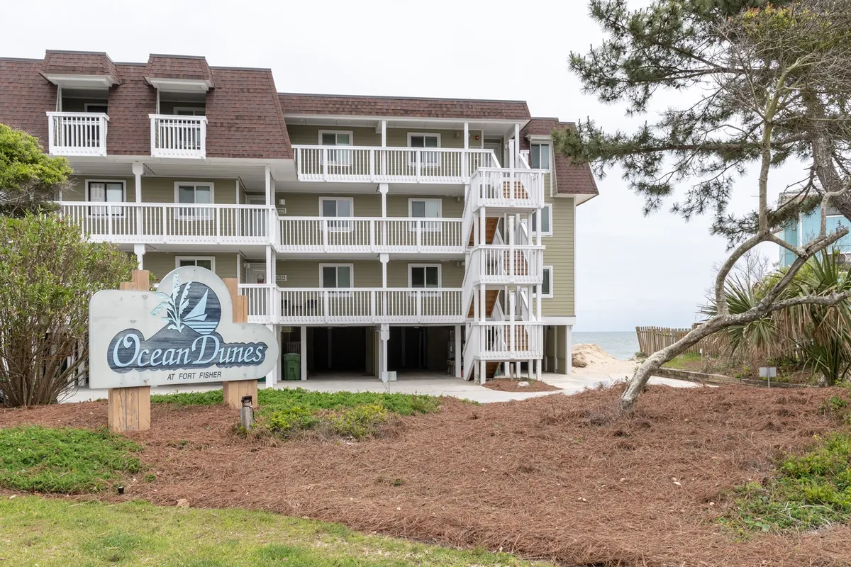 A welcoming exterior shot of the Ocean Dunes property, showing the building facade with balconies, an inviting sign, and stairs leading up to the units. The scene is tranquil, with a hint of ocean views and a sense of arrival for guests.