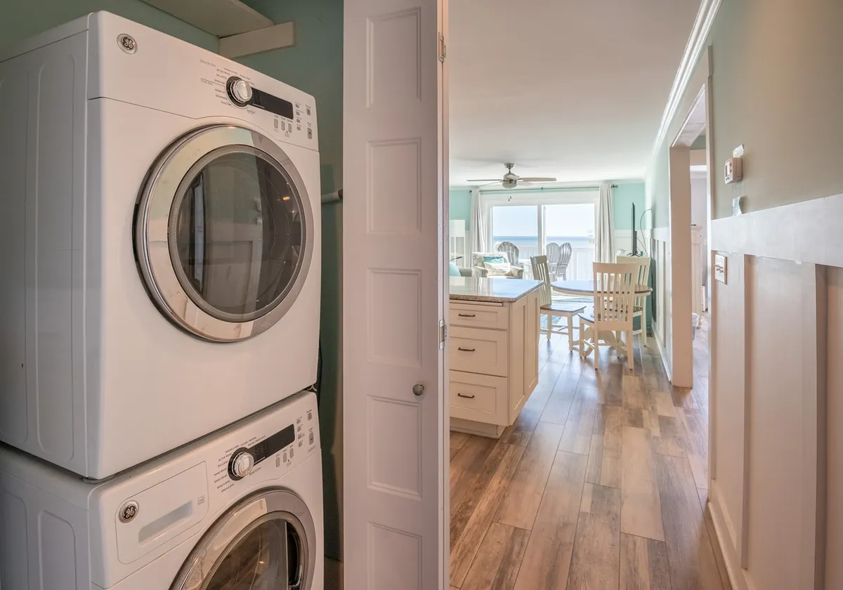 Shows a practical laundry area with a stacked washing machine and a view into a welcoming dining area through the doorway. The space feels functional yet inviting, suggesting ease for guests to handle laundry needs during their stay.