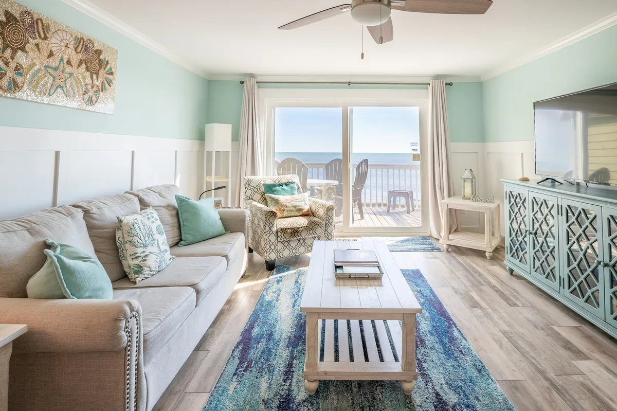 A bright, open living room with comfortable sofa and decorative pillows, a coffee table, large windows framing sweeping ocean views, and elegant wall art. The scene is filled with natural light, inviting relaxation and social gatherings, with easy access to the adjacent deck visible through the expansive windows.