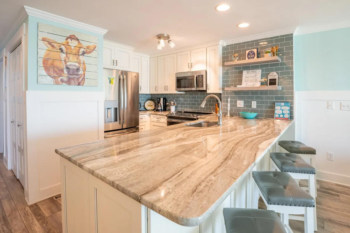 The kitchen is depicted with a granite island countertop, a playful cow portrait, and a stylish blue tile backsplash, creating a warm, inviting atmosphere perfect for cooking and gathering. The space appears fully equipped, aligning well with the description of a stocked kitchen.