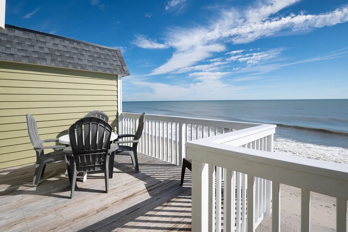 The image showcases a spacious deck with comfortable seating overlooking an expansive ocean view under a softly clouded sky. The serene atmosphere invites guests to relax, enjoy morning coffee, or gather for sunset watching. The proximity to the beach and the unobstructed coastal vista are immediately apparent, promising a tranquil seaside experience.