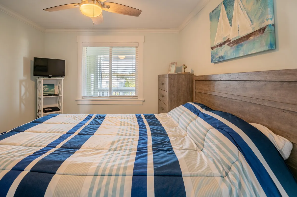 This image showcases a tranquil bedroom with a bed dressed in blue and white striped bedding, accented by wall art featuring sailboats. Natural light streams in through the window, and a TV is positioned on a stand, providing both relaxation and entertainment.