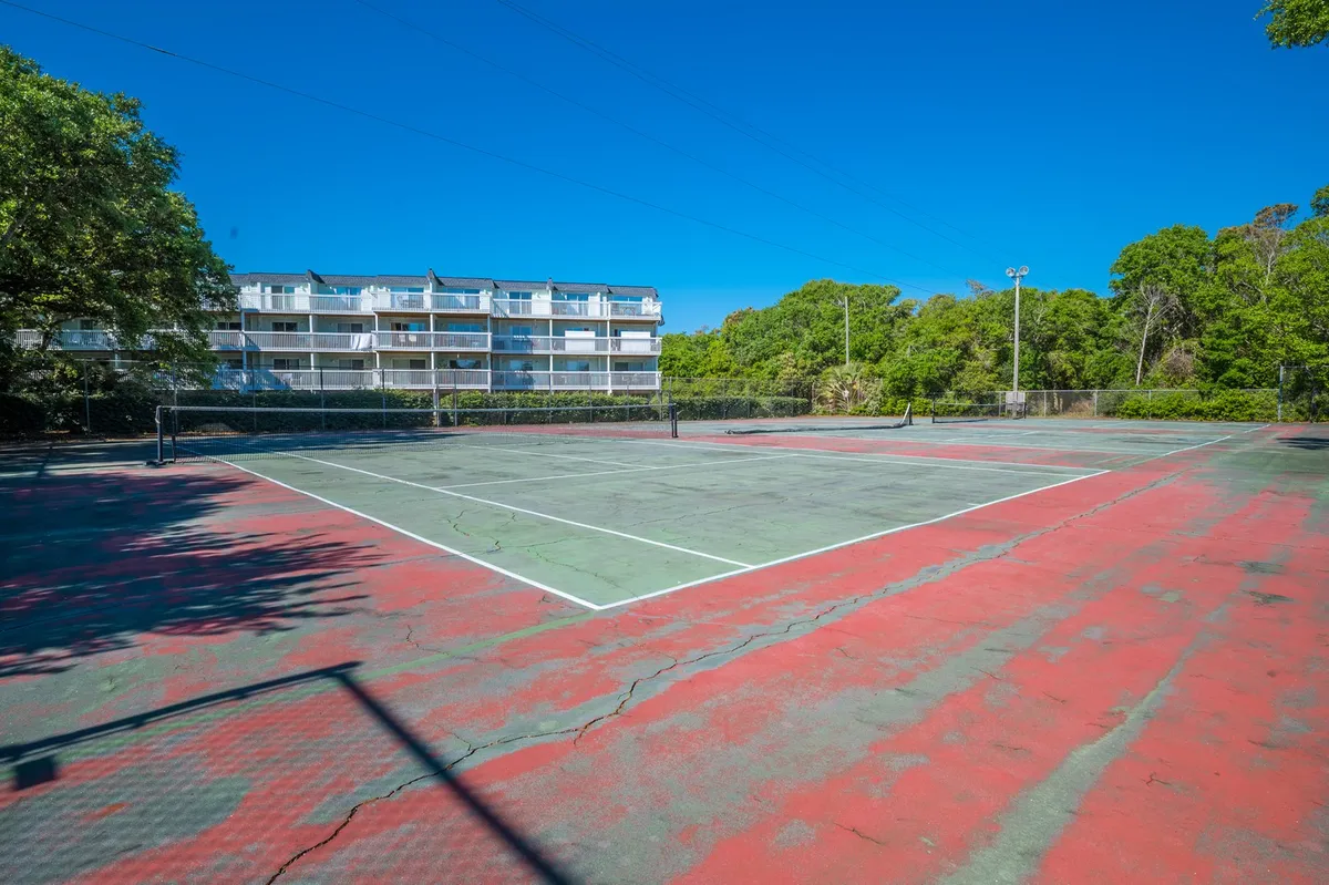A well-maintained outdoor tennis court bordered by greenery with an apartment building in the background. The open court invites guests for casual play or friendly matches under clear skies.